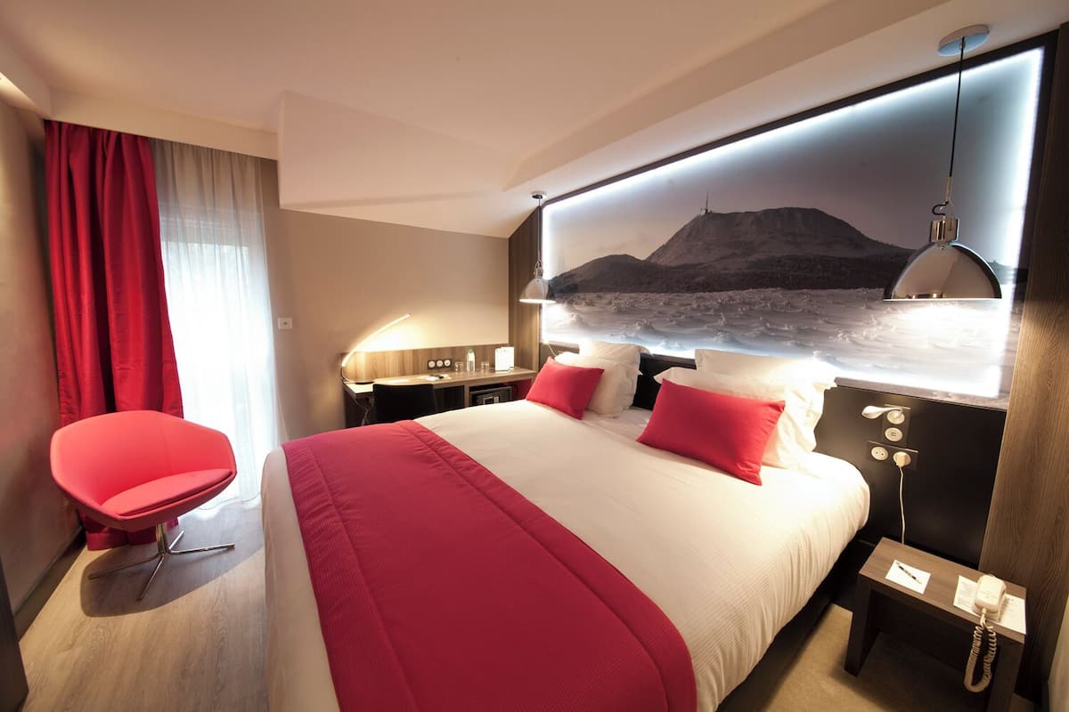 Hotelkamer met een groot bed, rood-wit beddengoed, een rode stoel, een bureau met een lamp en een wandtelevisie waarop een berglandschap te zien is.