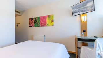 A hotel room with a bed, a desk, a chair, a radiator, and a painting on the wall.
