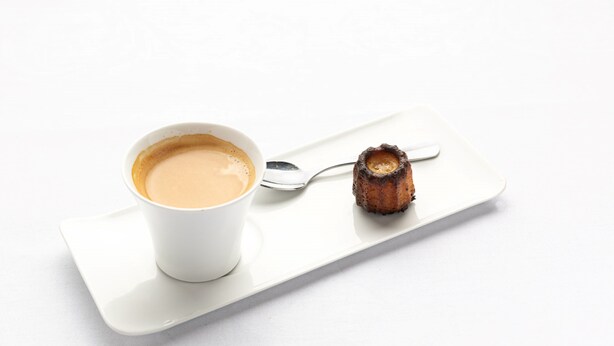 Une tasse de café blanc sur un plateau blanc, accompagnée d’une cuillère et d’une tranche de pain grillé brûlé.