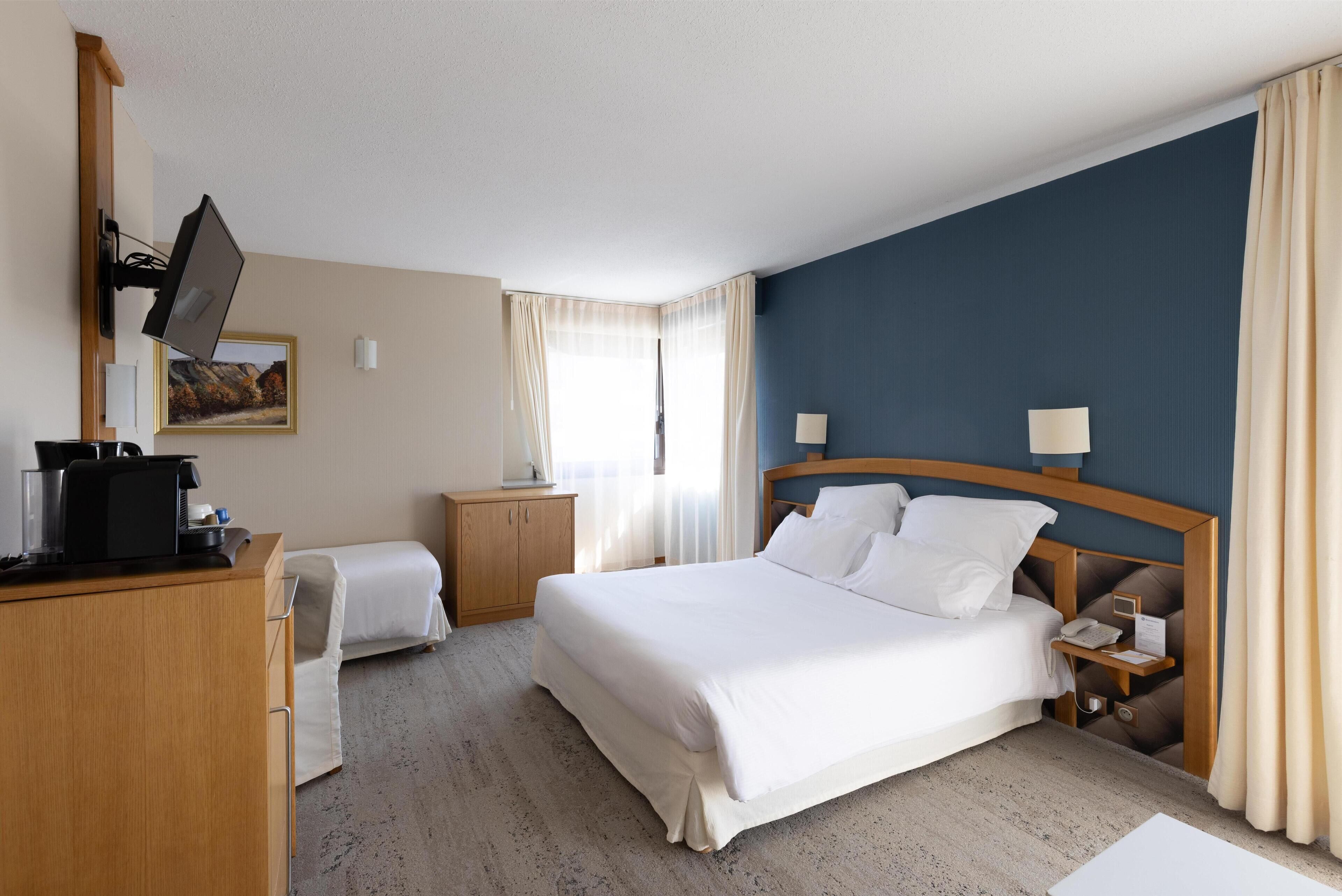 Ein Hotelzimmer mit einem großen Bett, Nachttischen, einem Schreibtisch mit Computer und einem an der Wand montierten Fernseher.