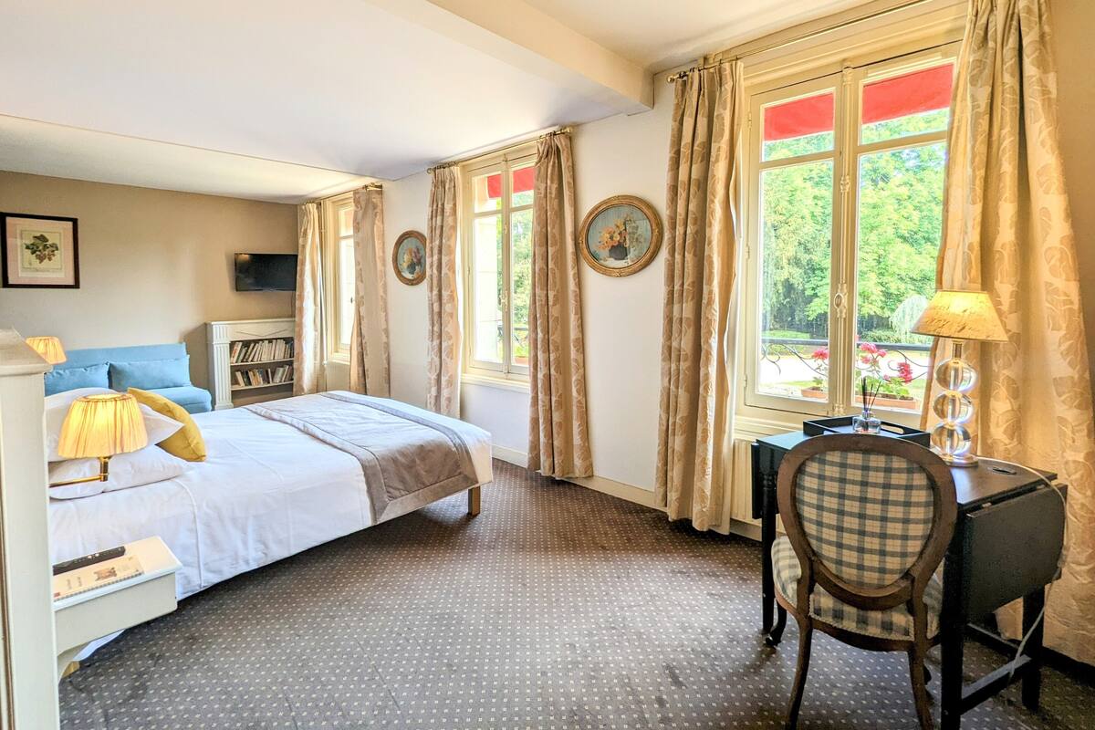 Habitación de hotel con cama, escritorio, silla, estantería y ventana con cortinas.