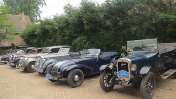 A row of vintage cars parked on a gravel surface, surrounded by trees and a building with a tiled roof.