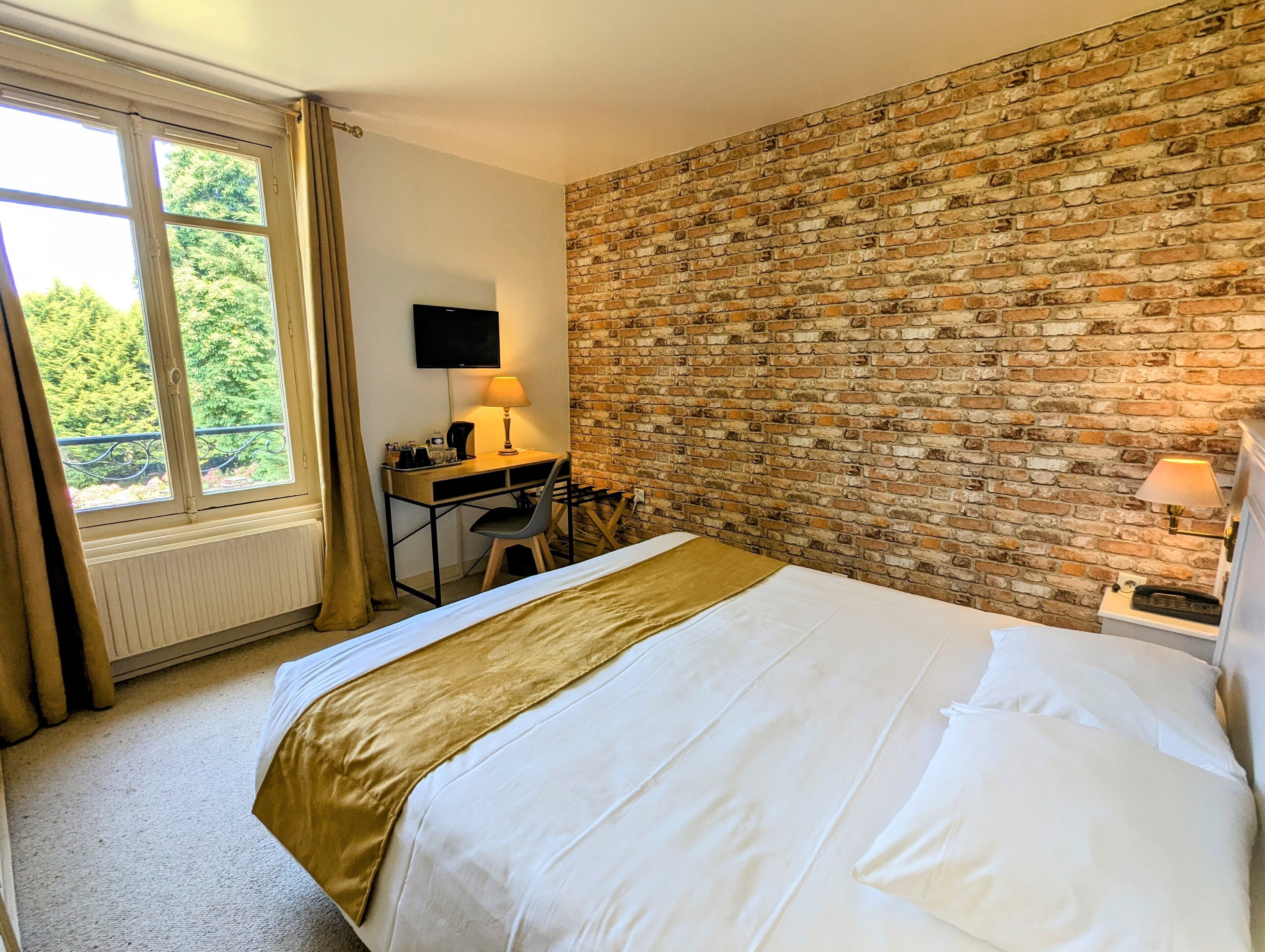 A hotel room with a brick wall, a bed with white linens, a desk with a lamp, and a window with curtains.