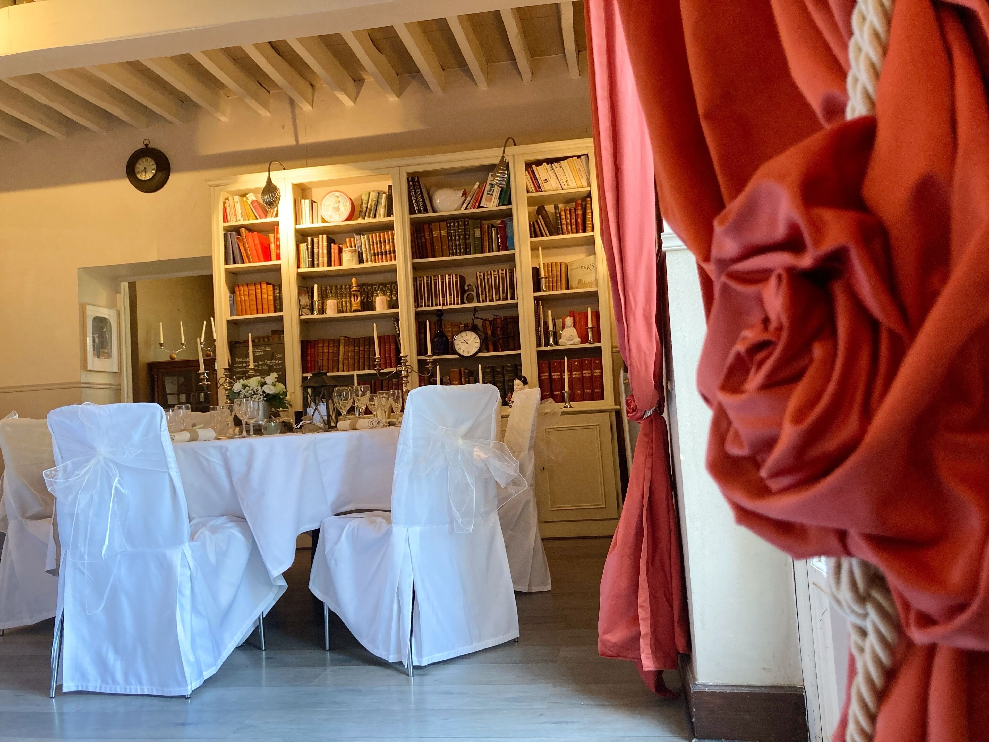 A room with a dining table set for six, surrounded by white chairs with sashes, a bookshelf filled with books, and a clock on the wall.