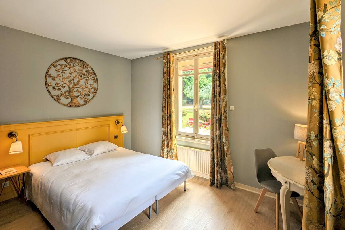 Un dormitorio con cama, un adorno circular en la pared, una ventana con cortinas y una pequeña mesa con silla.
