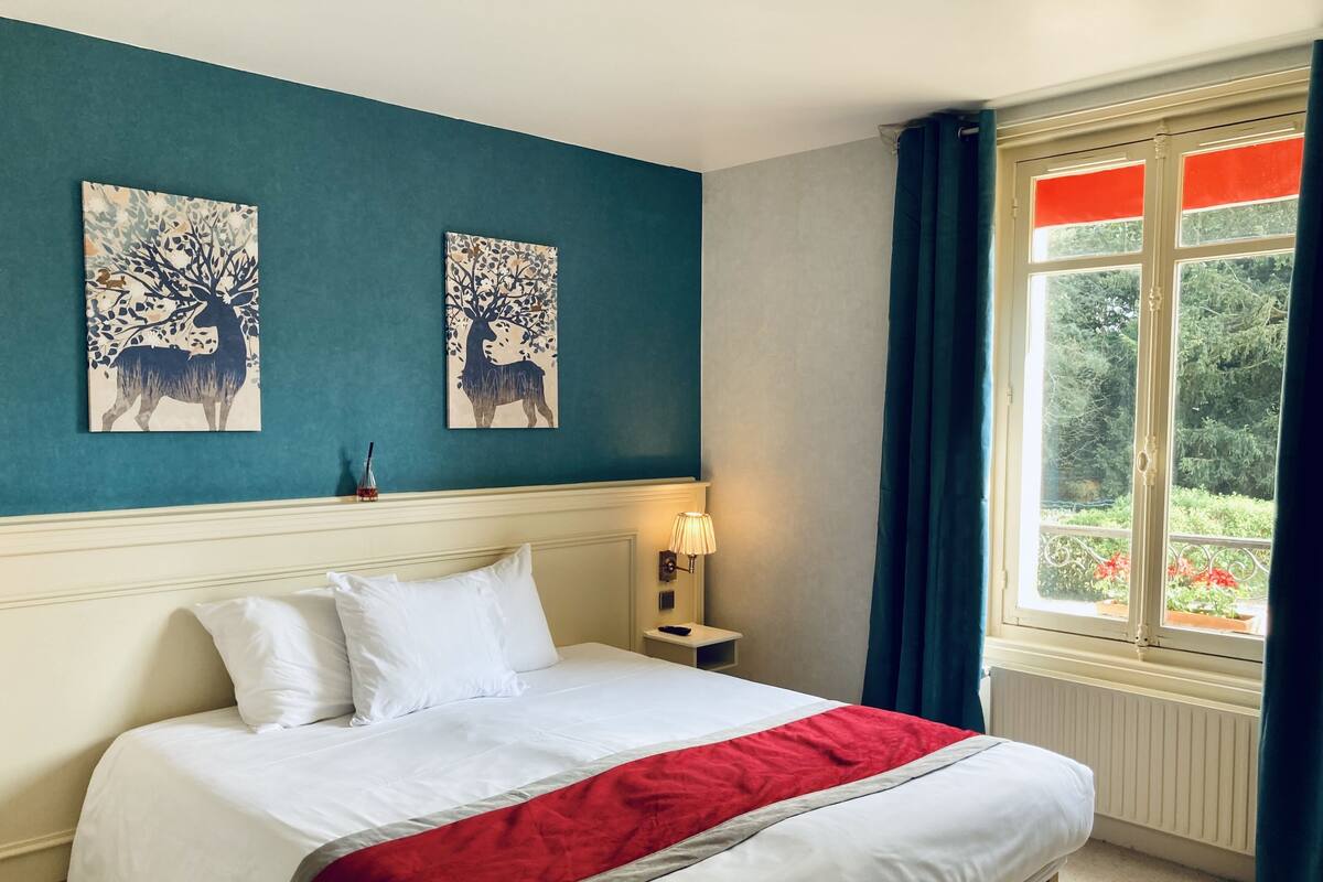 Habitación de hotel con cama, dos cuadros decorativos, una mesita de noche, lámpara y ventana con cortinas.