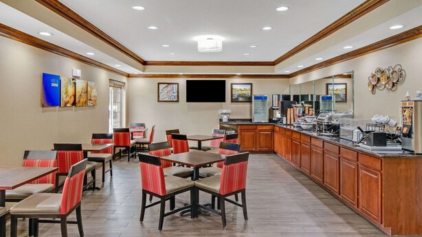A hotel dining area with multiple tables and chairs, a kitchen counter with coffee machines, and a flat-screen TV mounted on the wall. - Comfort Inn Henderson - Evansville South (Henderson)