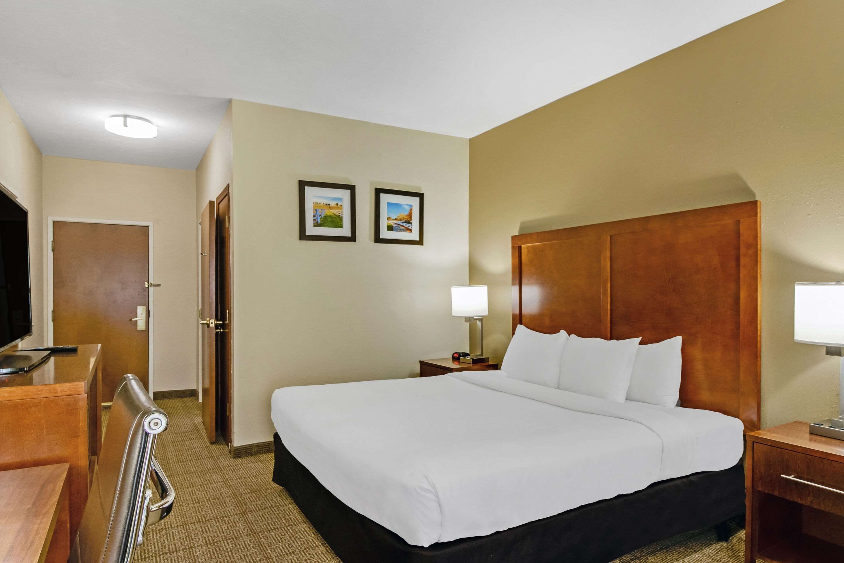A hotel room with a large bed, a desk with a chair, a TV, and two framed pictures on the wall.