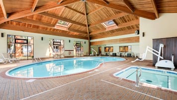 Una piscina interior con dos zonas separadas, rodeada de sillones de descanso y mesas, bajo un techo de madera con tragaluces.