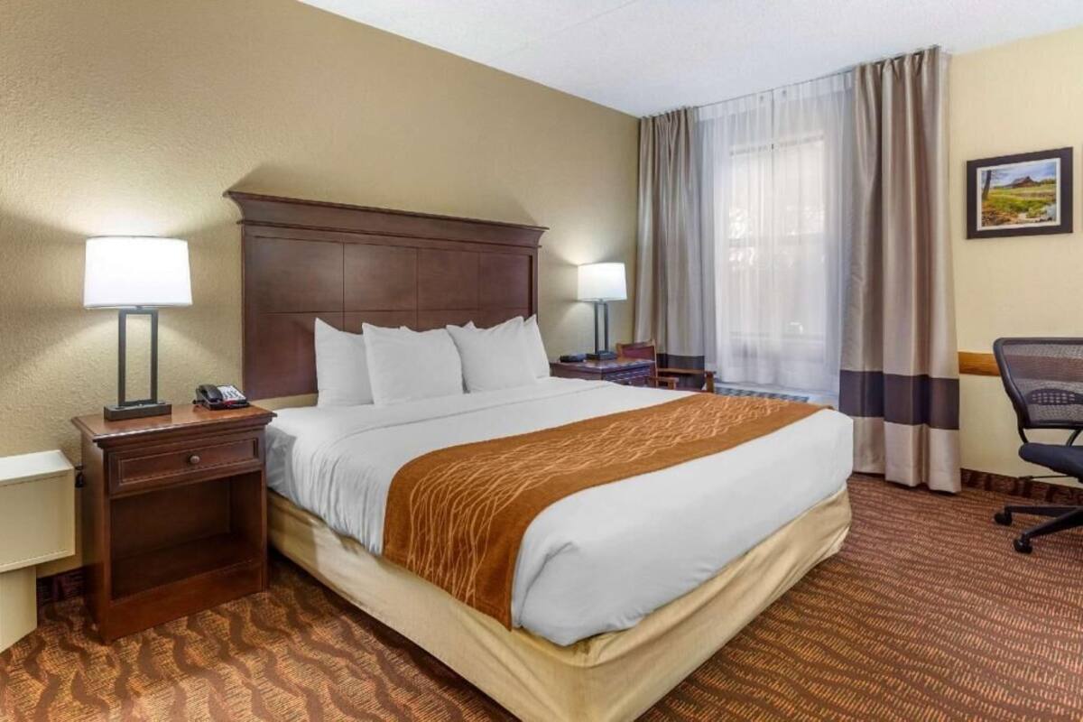 A hotel room with a large bed, two bedside tables with lamps, a desk with a chair, and a window with curtains.