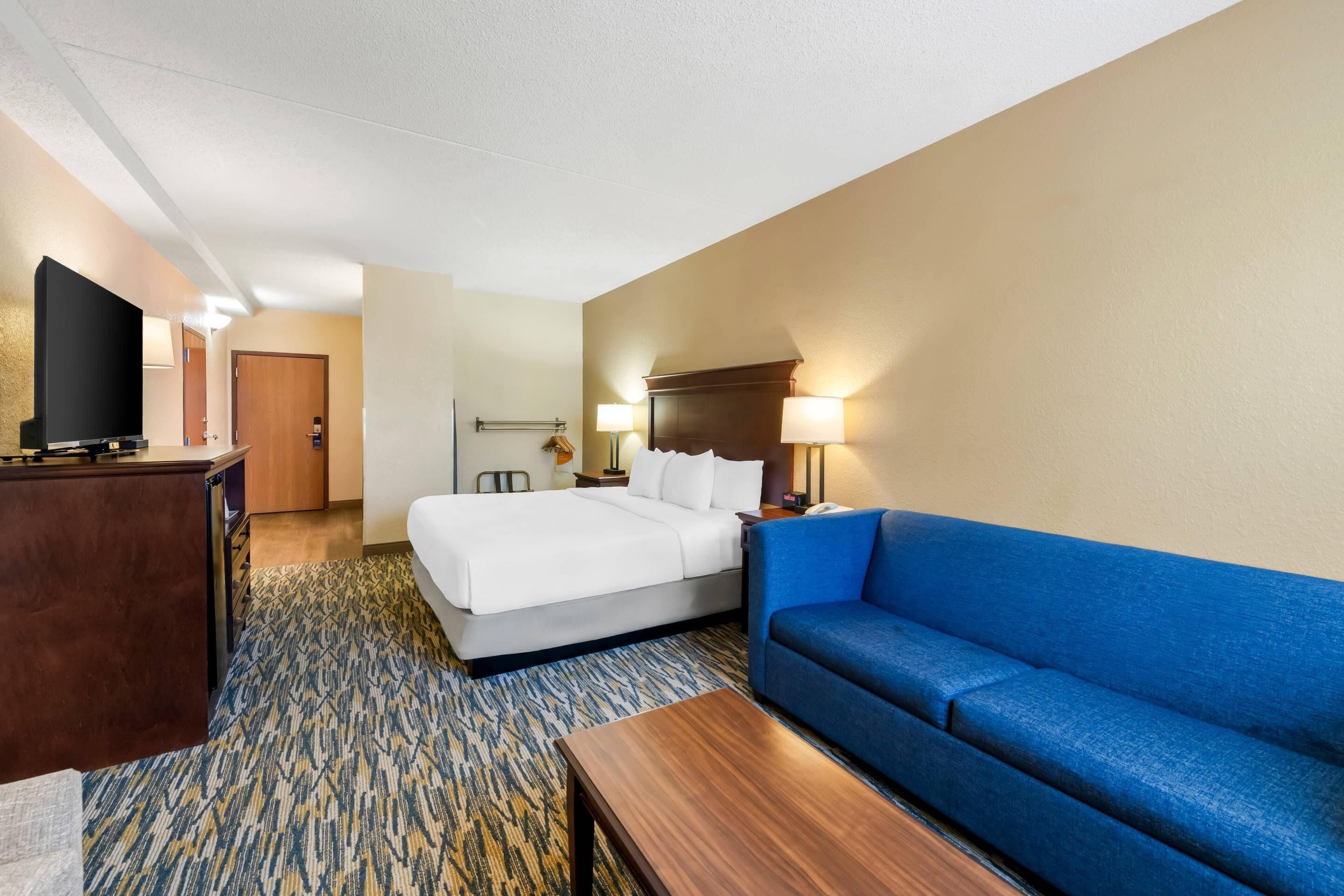 A hotel room with a bed, a blue sofa, a wooden desk, a television, and a lamp.