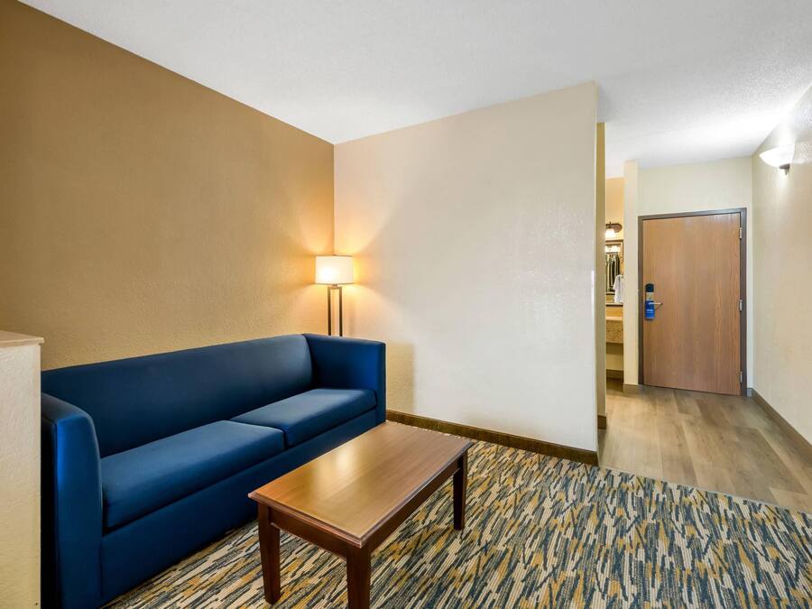 A hotel room with a blue sofa, a wooden coffee table, a patterned carpet, and a door leading to a hallway.