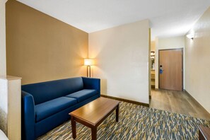 Hotellihuoneessa on sininen sohva, puinen sohvapöytä, kuviollinen matto ja ovi, joka johtaa käytävään.