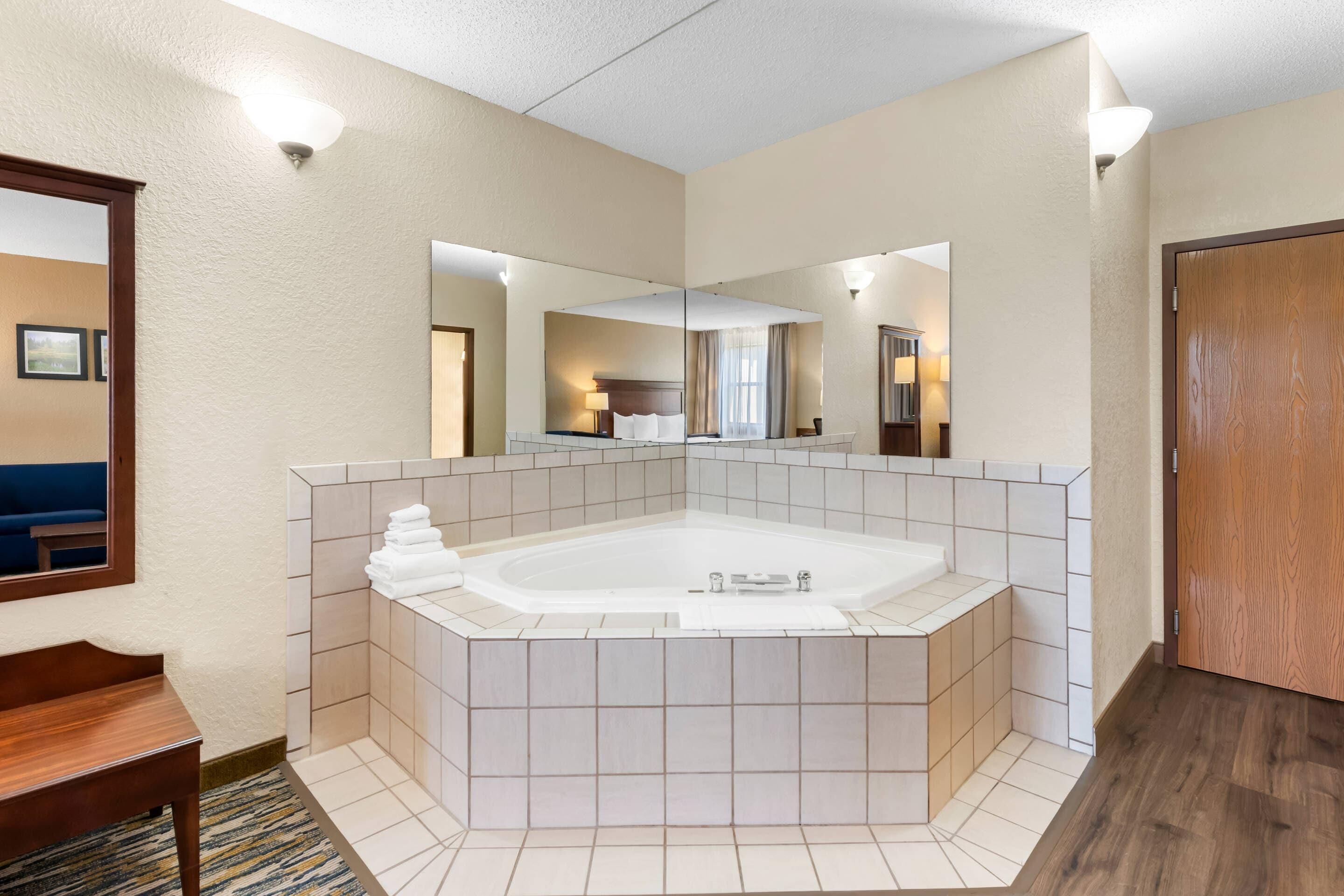 A hotel bathroom with a large bathtub, a mirror, and a wooden bench.