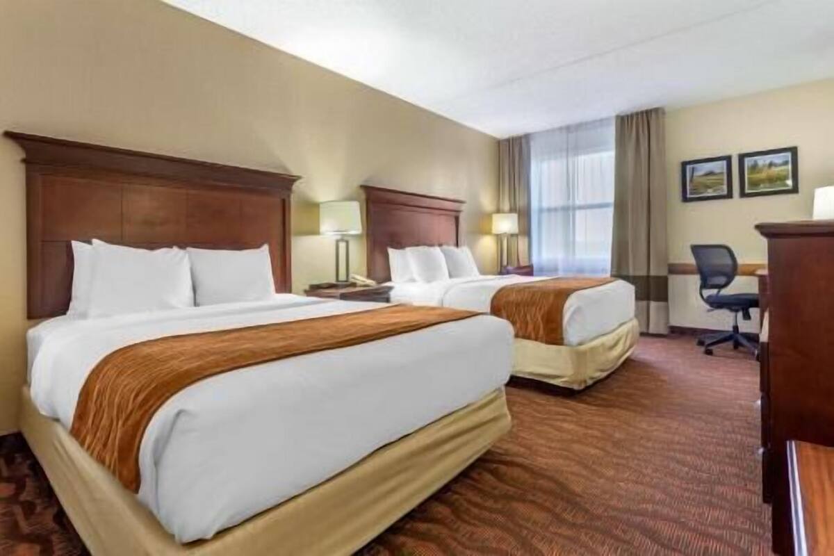 A hotel room with two beds, a desk, a chair, and a lamp.