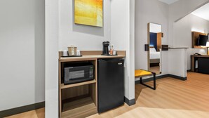 Habitación de hotel compacta con kitchenette, microondas, refrigerador, una mesita con una silla, un cuadro en la pared y televisor sobre un mueble.