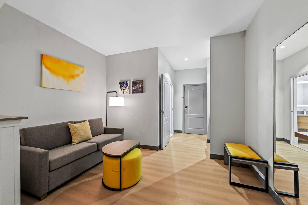Een moderne hotelkamer met een grijze bank, een geel poufje, een klein tafeltje en een grote spiegel.
