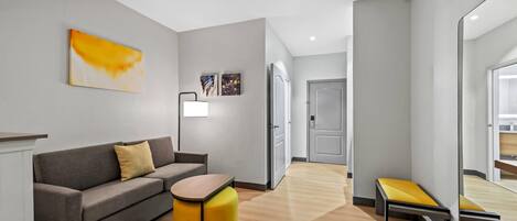 A modern hotel room with a grey sofa, a yellow ottoman, a small table, and a large mirror.