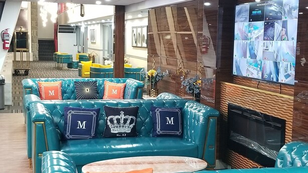 A hotel lobby with teal sofas, a wooden floor, and a large screen displaying multiple rooms.