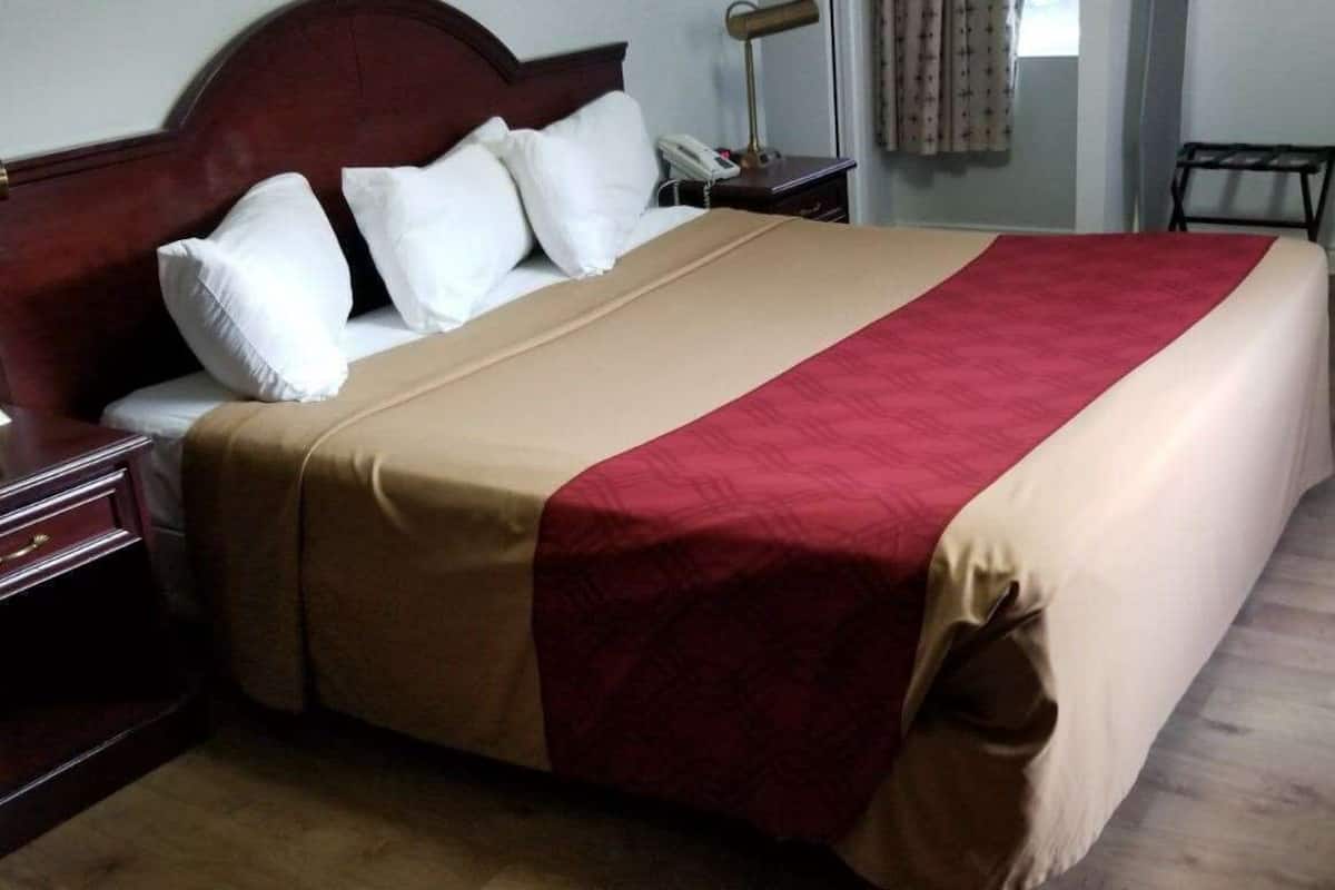 A neatly made bed with a red and beige bedspread, a wooden headboard, a bedside table with a lamp, and a chair.