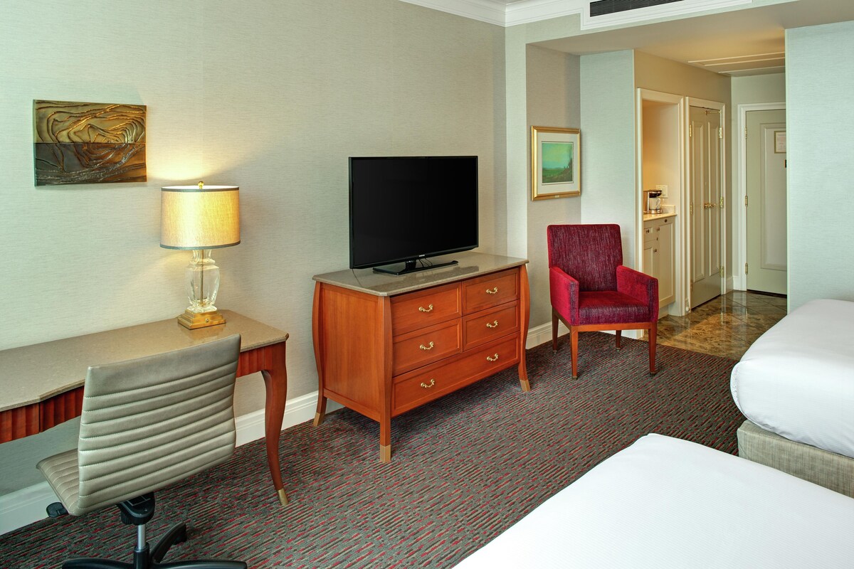 Una habitación de hotel con televisión de pantalla plana, un escritorio con lámpara, una silla roja, una cama y suelo alfombrado.