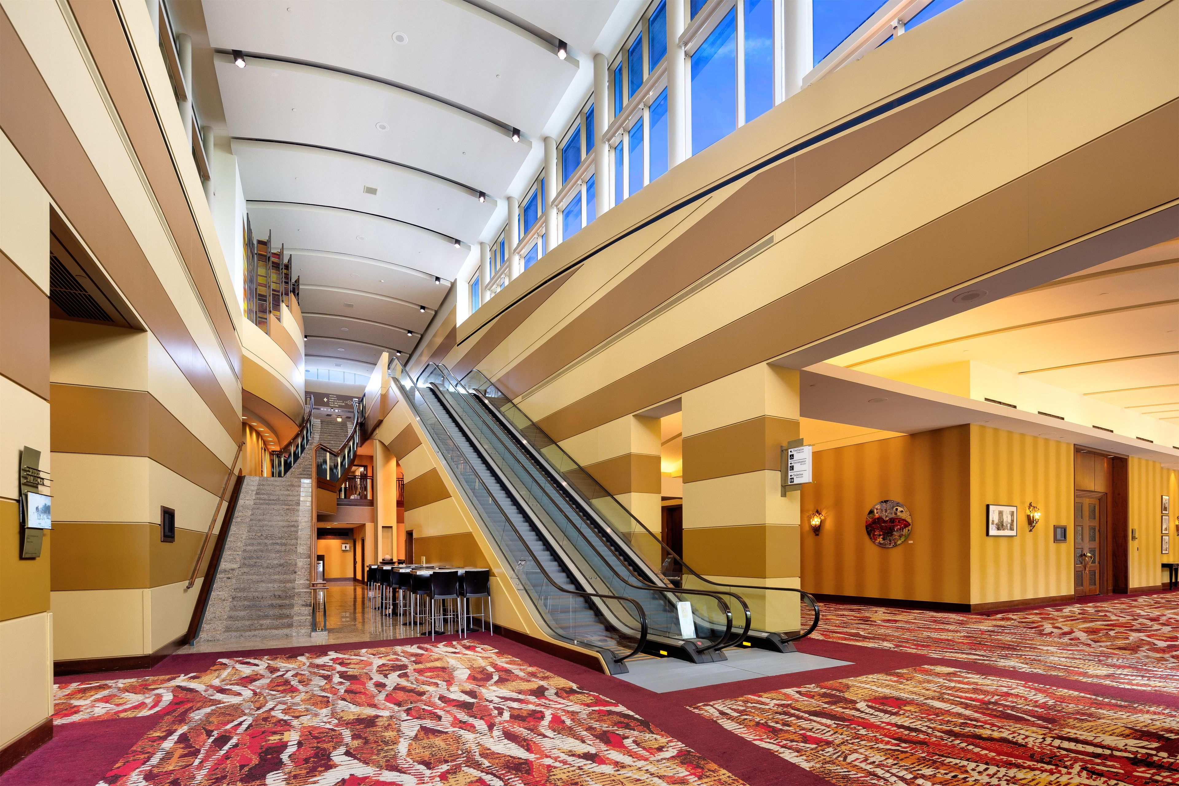 Un vestíbulo de hotel con una escalera mecánica central, una escalera y suelo alfombrado.
