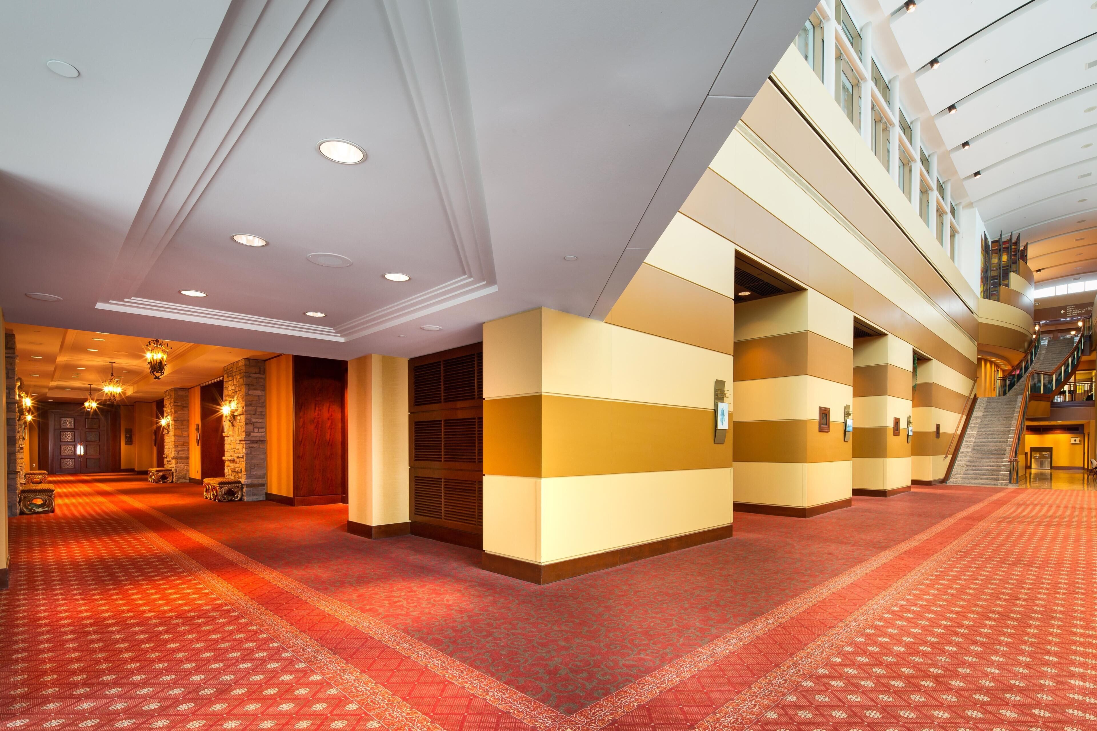 Un amplio vestíbulo de hotel con alfombra roja, varias puertas y una escalera que conduce a pisos superiores.
