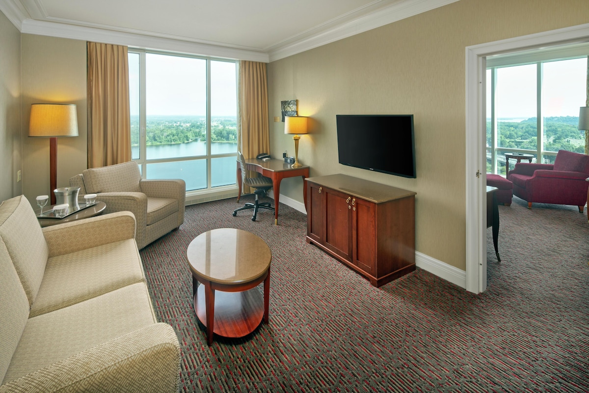 Habitación de hotel con sofá, una mesita, escritorio con lámpara, televisor y vista al lago.