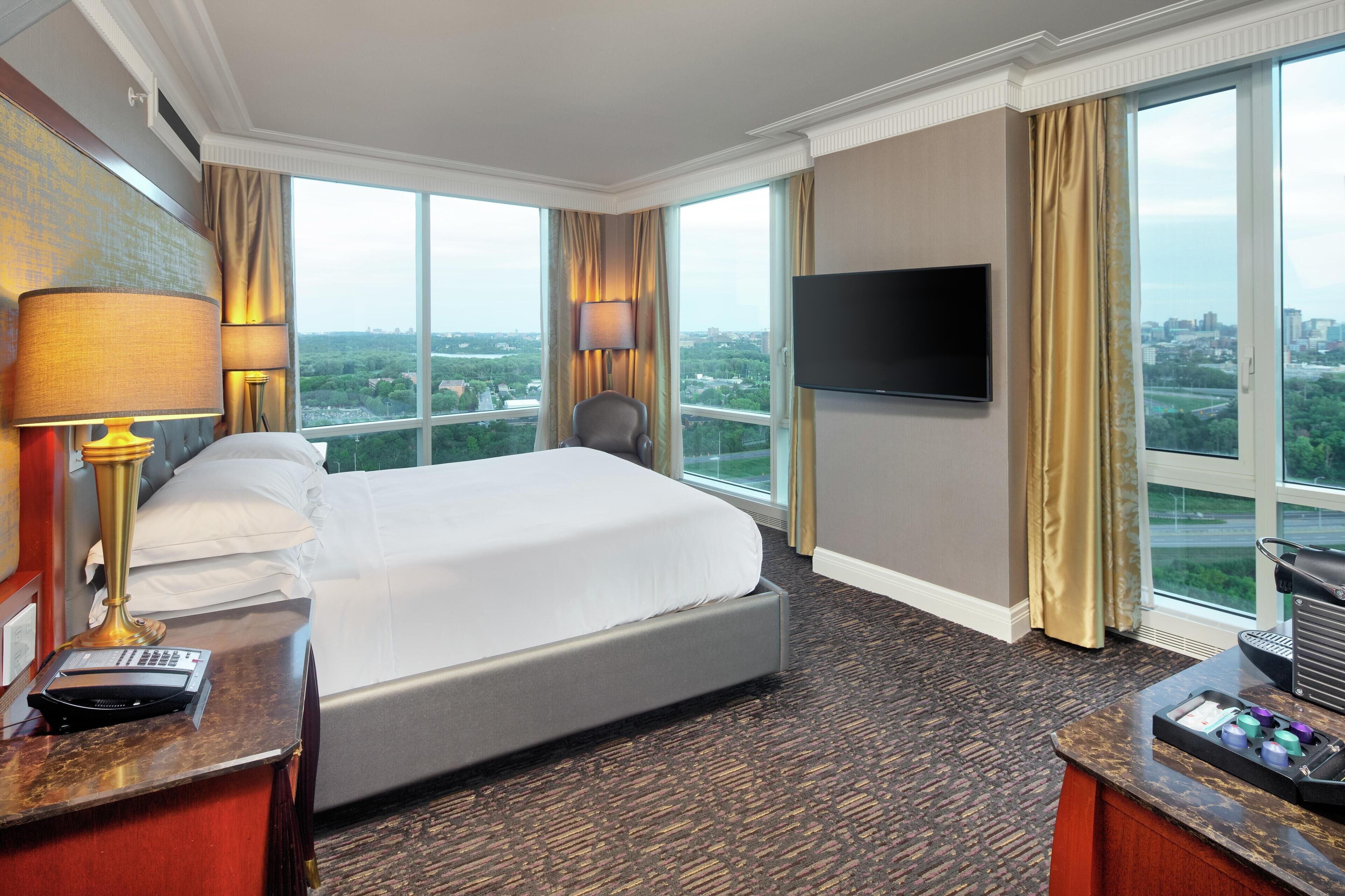 Een hotelkamer met een groot bed, een bureau met lamp, een televisie en uitzicht op de stad.