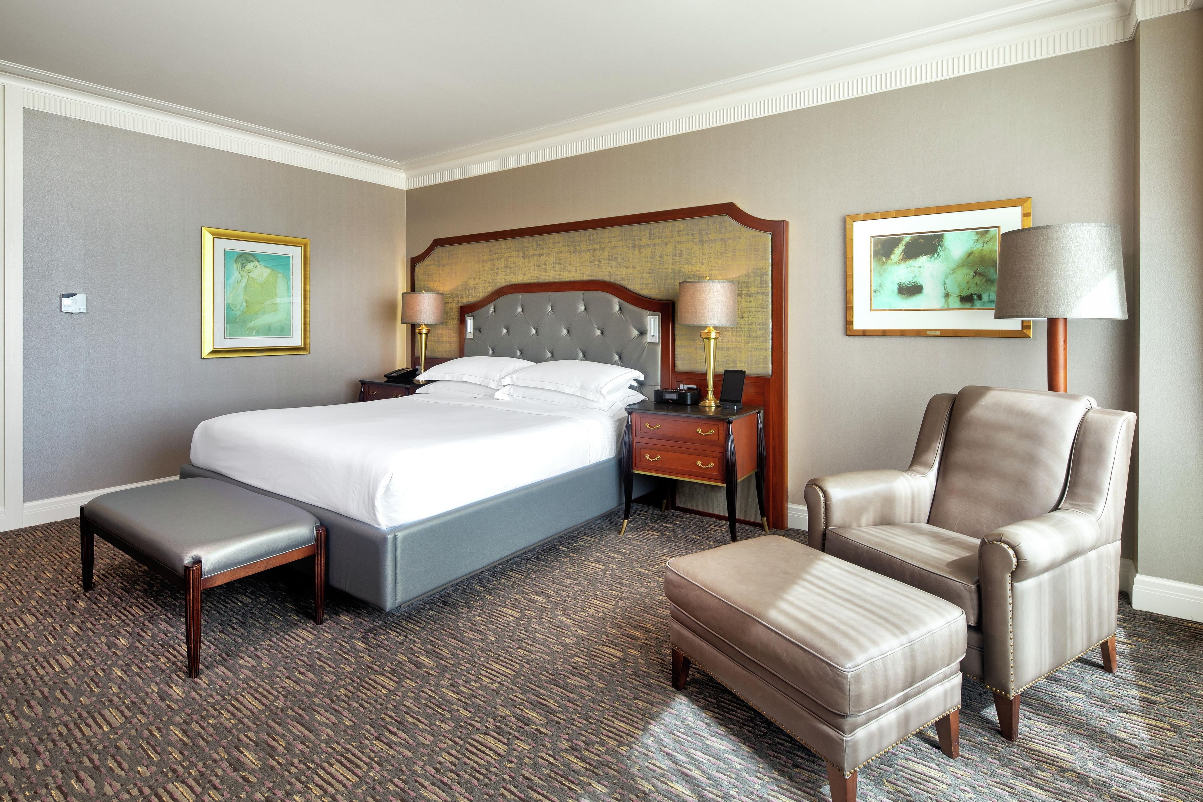 Quarto de hotel com uma cama grande, duas mesas de cabeceira, uma poltrona e um banquinho, além de duas fotografias emolduradas nas paredes.