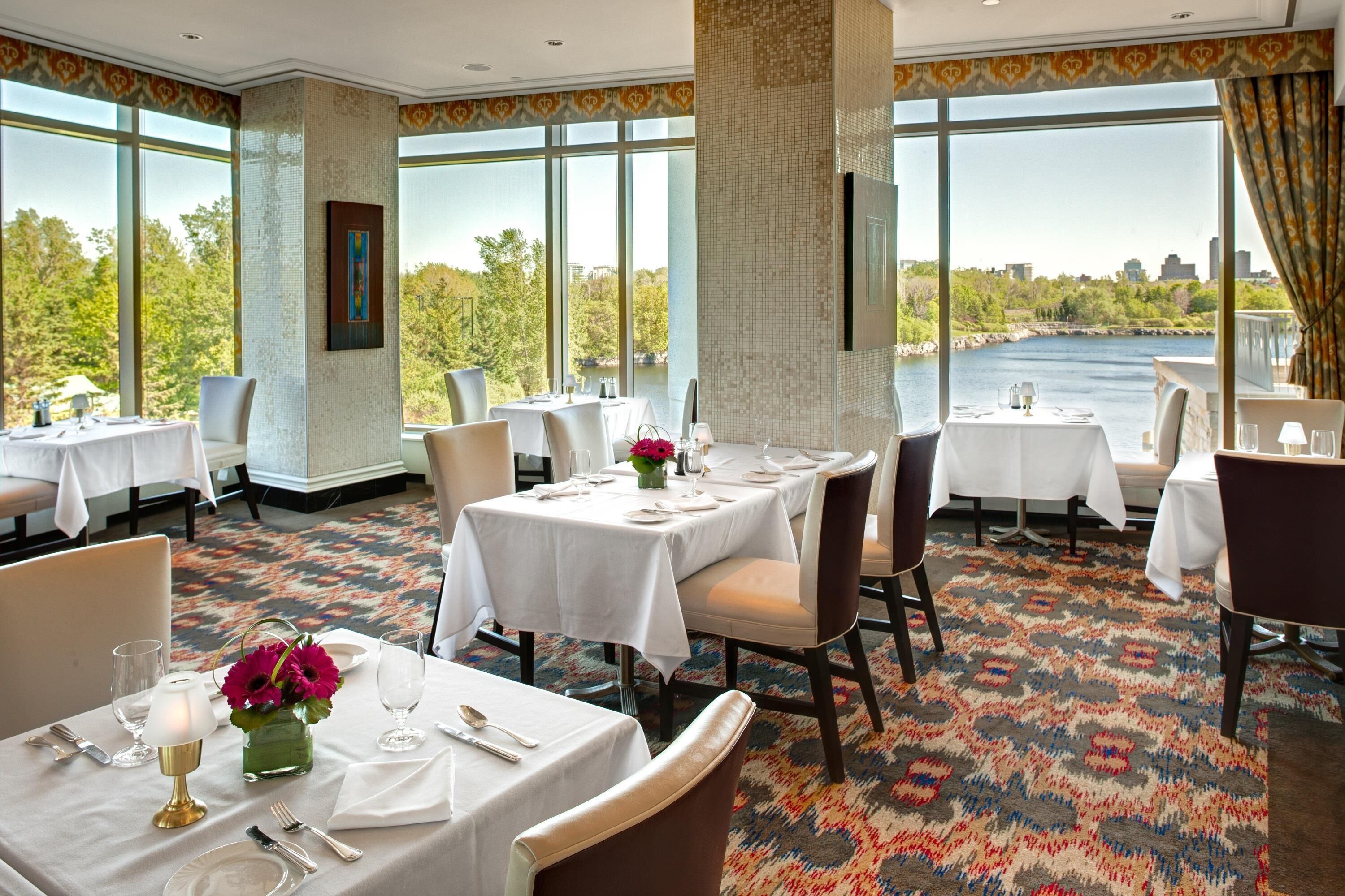 A dining area with tables set for four, large windows with a view of a body of water and greenery, and a patterned carpet.