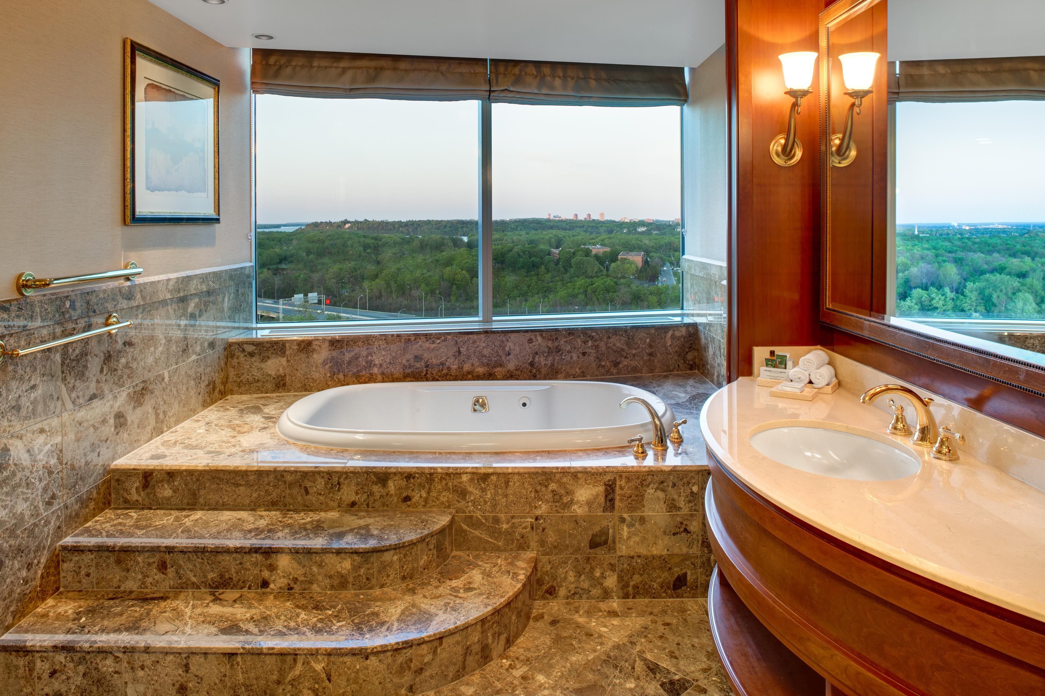 Une salle de bain dotée d’une grande baignoire, d’un lavabo séparé et offrant une vue sur une zone boisée grâce à de grandes fenêtres.