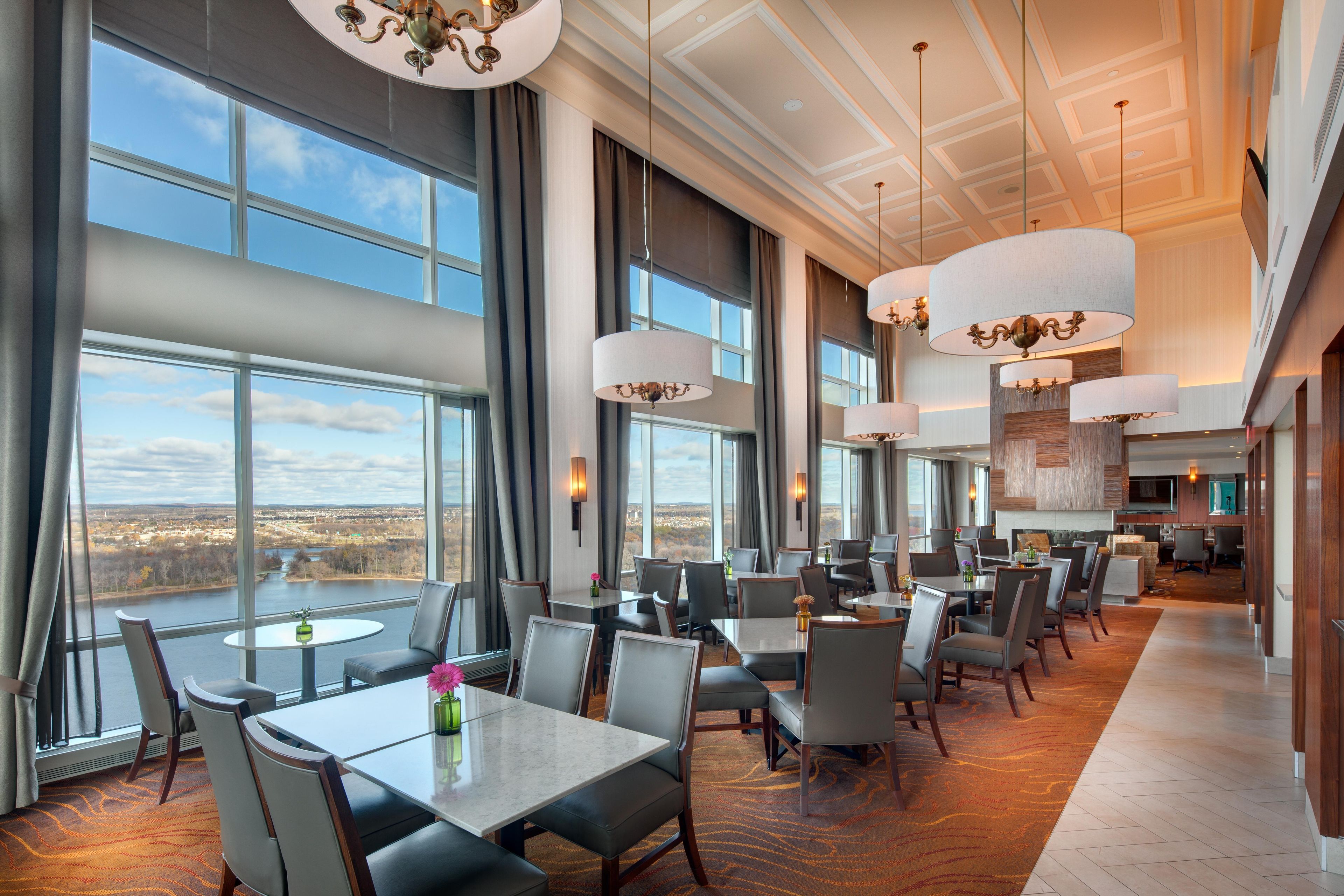 A spacious hotel dining area with large windows, multiple tables, and elegant chandeliers.