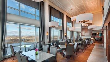 A spacious hotel dining area with large windows, multiple tables, and elegant chandeliers.
