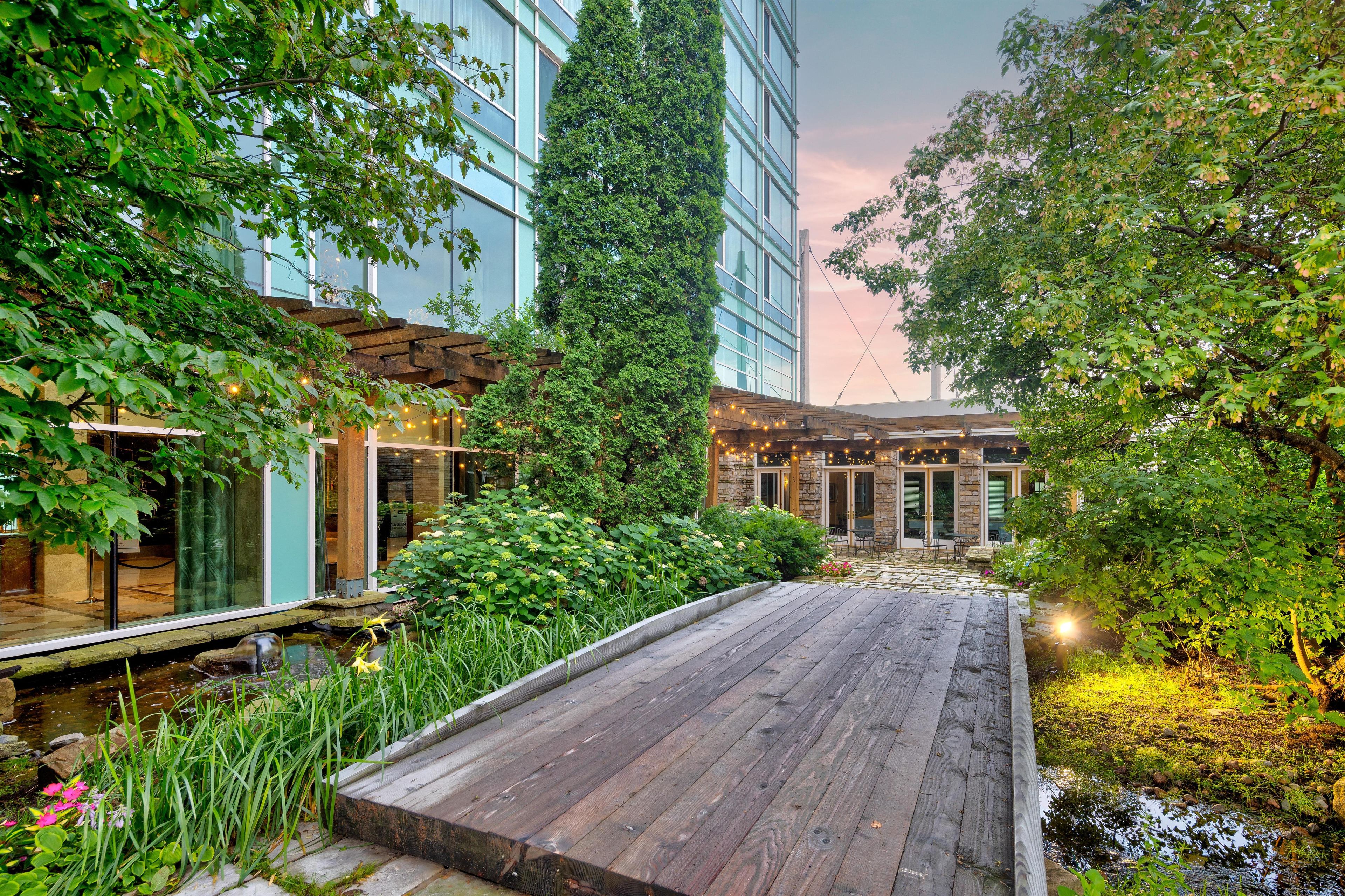 Una terraza de madera con un estanque, rodeada de abundante vegetación y edificios modernos.