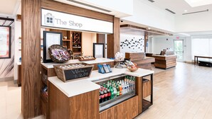 A reception area with a wooden counter, a refrigerated display of beverages, and a sign reading "The Shop". - Hilton Garden Inn Fairfax (Fairfax)
