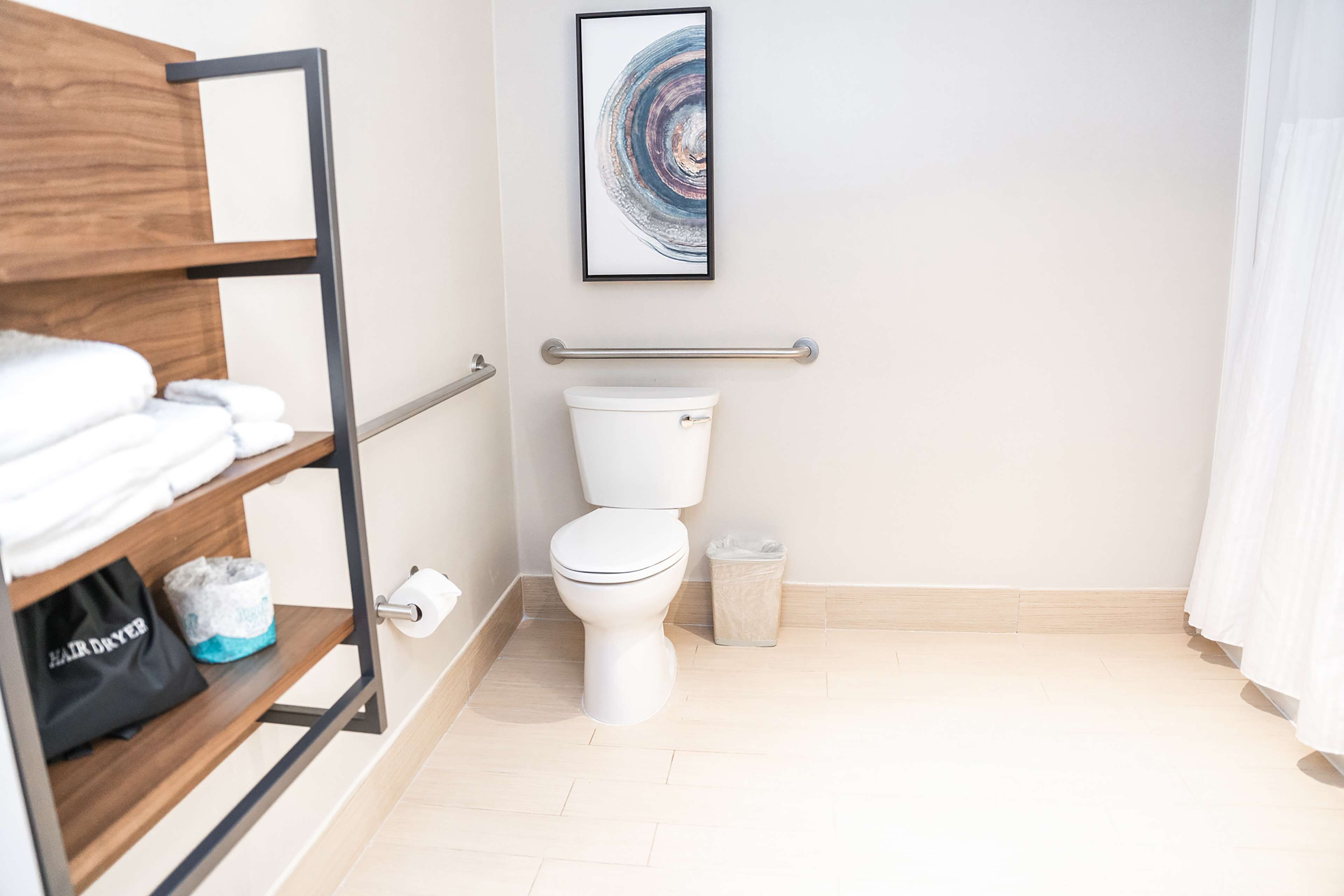 Ein modernes Badezimmer mit Toilette, einer Handstange, einem Ablagefach mit Handtüchern, einem Mülleimer und einem gerahmten Kunstwerk an der Wand.