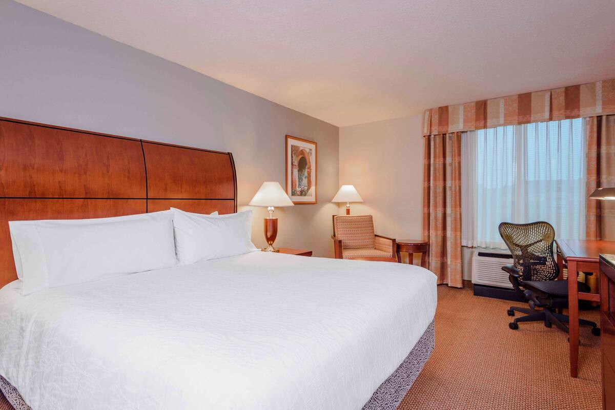 A hotel room with a large bed, two bedside lamps, a chair, a desk, and a window with curtains.
