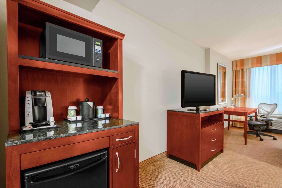A hotel room with a kitchenette, including a microwave, coffee maker, and refrigerator. There is a television on a wooden stand and a desk with a chair.