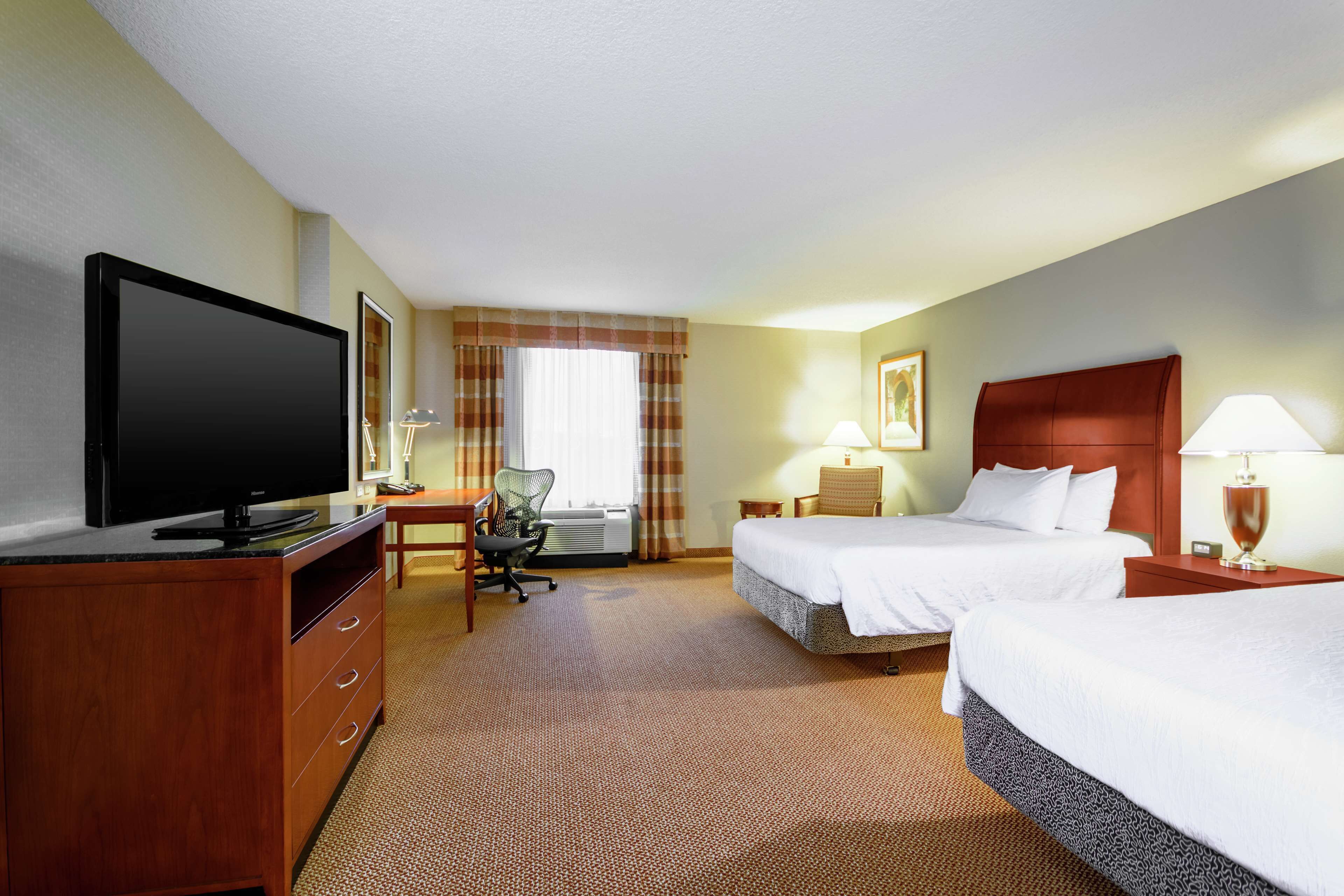 A hotel room with two beds, a TV, a desk with a chair, and a window with curtains.