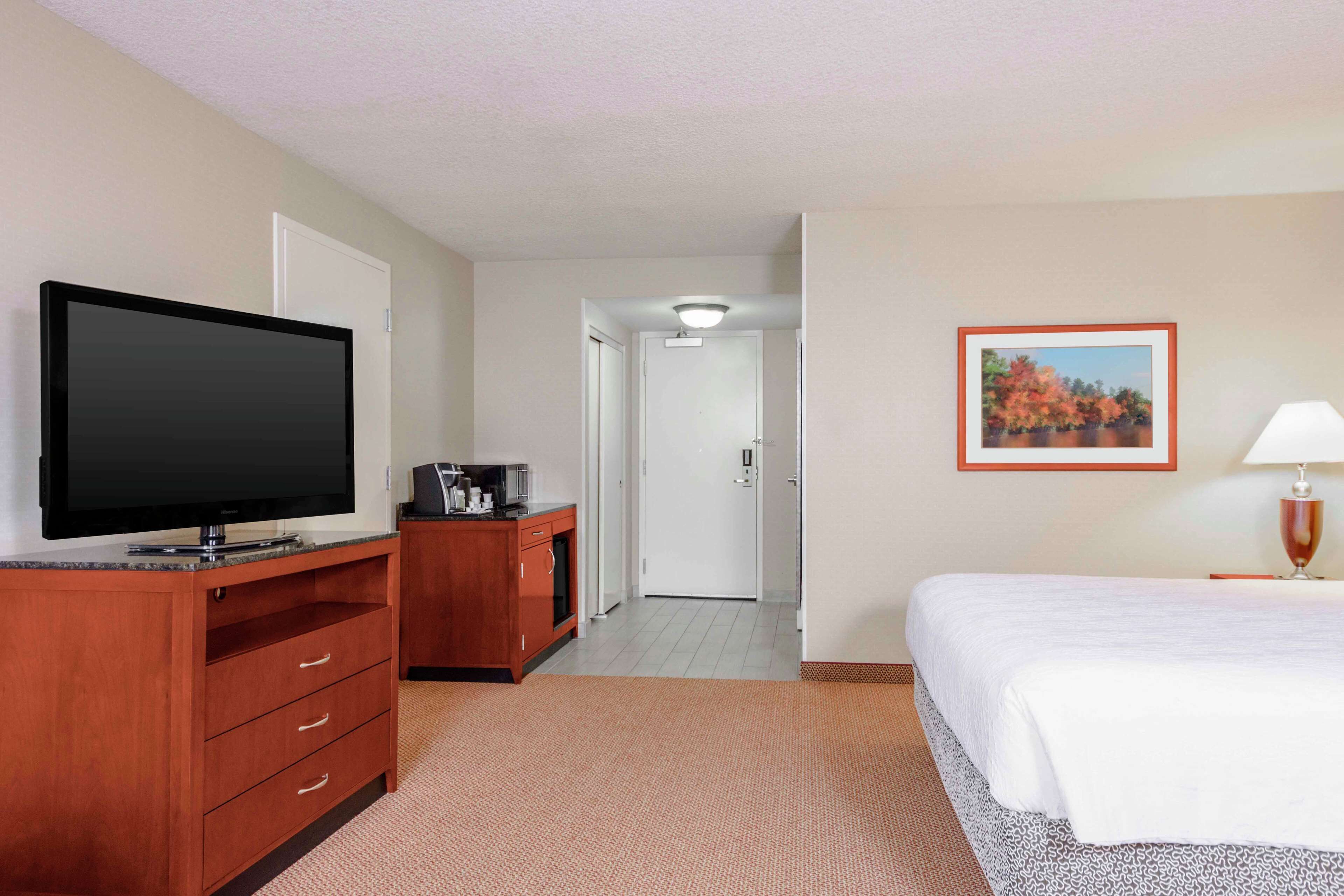 A hotel room with a flat-screen TV, a wooden dresser, a bed, a framed picture on the wall, and a lamp.