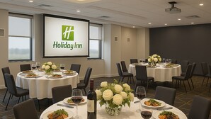 A conference room with multiple round tables set for dining, each with white tablecloths, chairs, and floral centerpieces. A large screen displays the Holiday Inn logo. - Holiday Inn & Suites Mississauga West - Meadowvale (Mississauga)