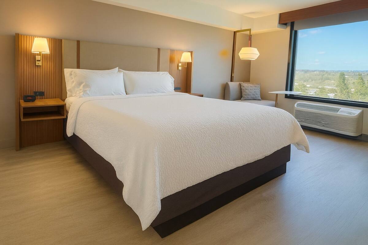 A modern hotel room with a large bed, a nightstand with a lamp, a chair, and a large window with a view.