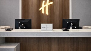 A reception desk with a wooden panel background and a large illuminated 'H' sign. There are two monitors on either side of the desk, and a sign in front that reads 'IHG ONE REWARDS'. - Holiday Inn & Suites Mississauga West - Meadowvale (Mississauga)