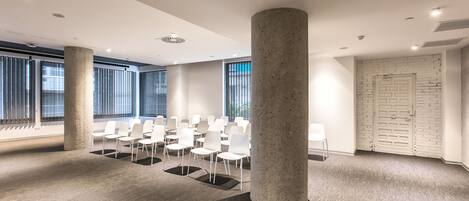 Un salón de conferencias con sillas blancas dispuestas en filas, ventanas grandes con persianas, una pared de ladrillo blanco y una puerta con diseño de cuadrícula.
