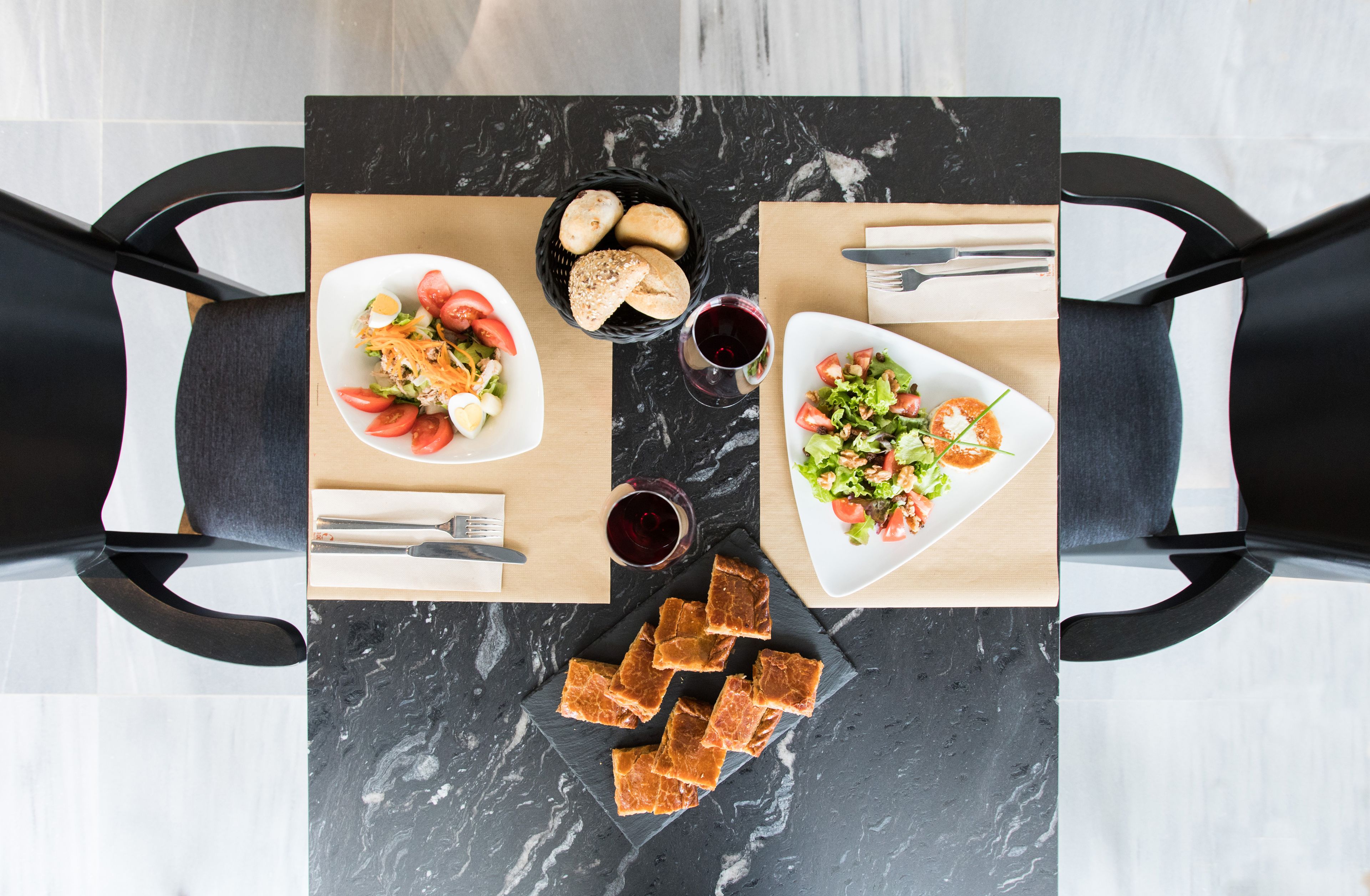 A dining table with two plates of salad, a basket of bread, and two glasses of red wine.