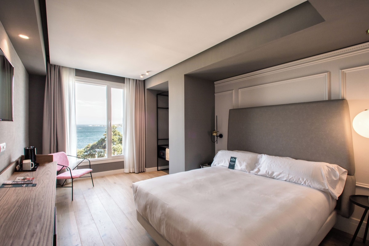 A modern hotel room with a large bed, a desk with a chair, a television, and a view of the ocean.