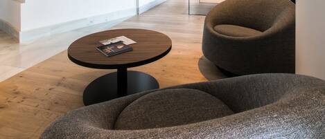 A modern lobby with a round wooden table, a grey armchair, and large potted plants.