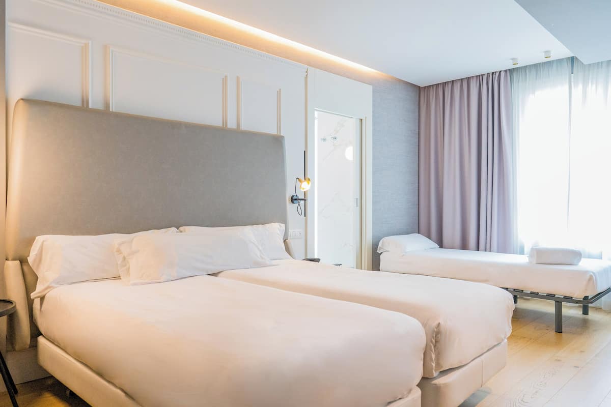 Hotellihuone, jossa on kaksi sänkyä, yöpöytä, lamppu, ikkuna verhoilla ja ovi.