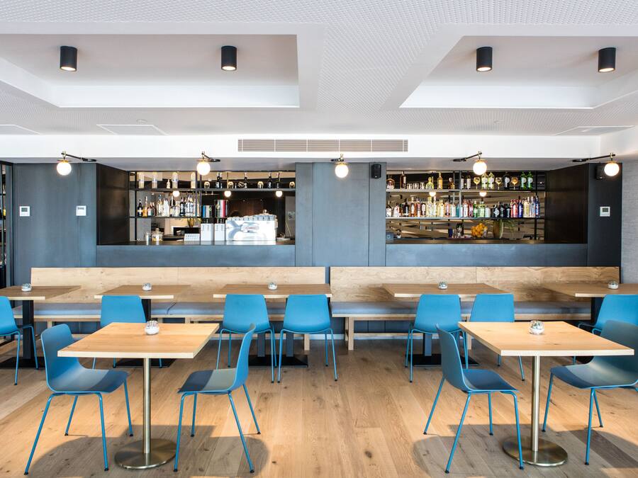 Un restaurante moderno con mesas de madera y sillas azules, una zona de bar con estanterías llenas de botellas y un área de asientos con bancos.